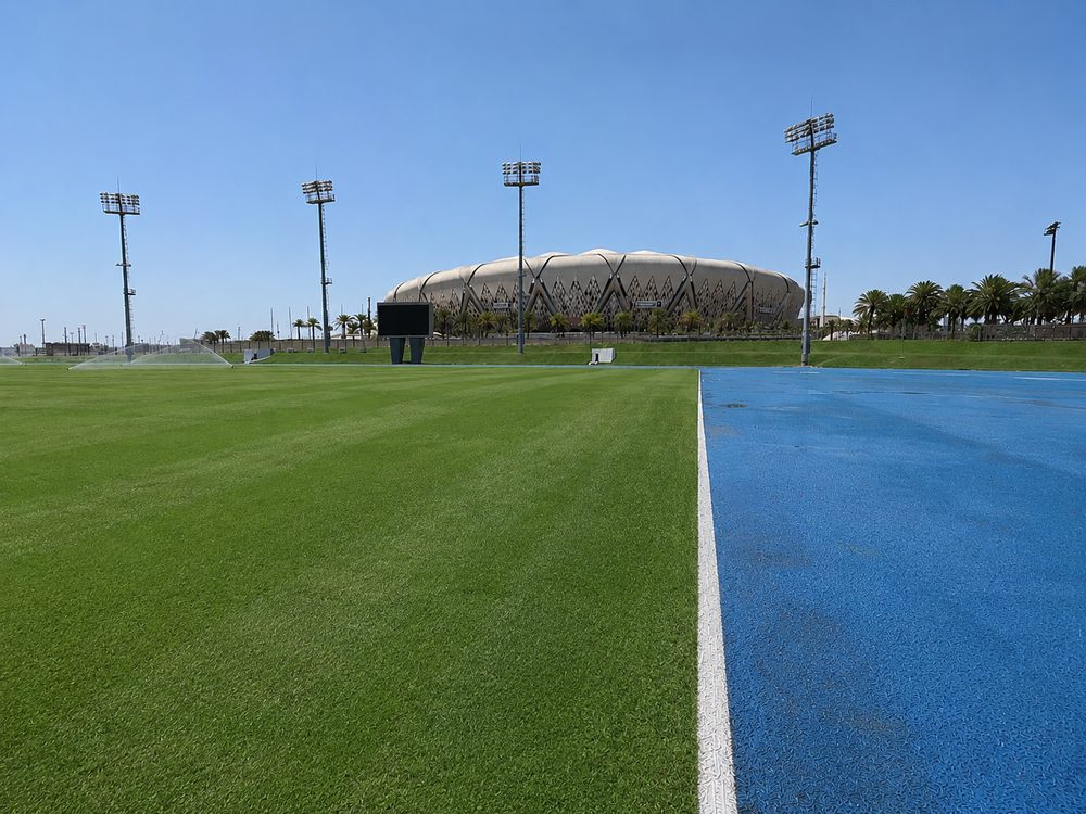 A grass training pitch meeting the blue edge of the athletics track, main stadium beyond
